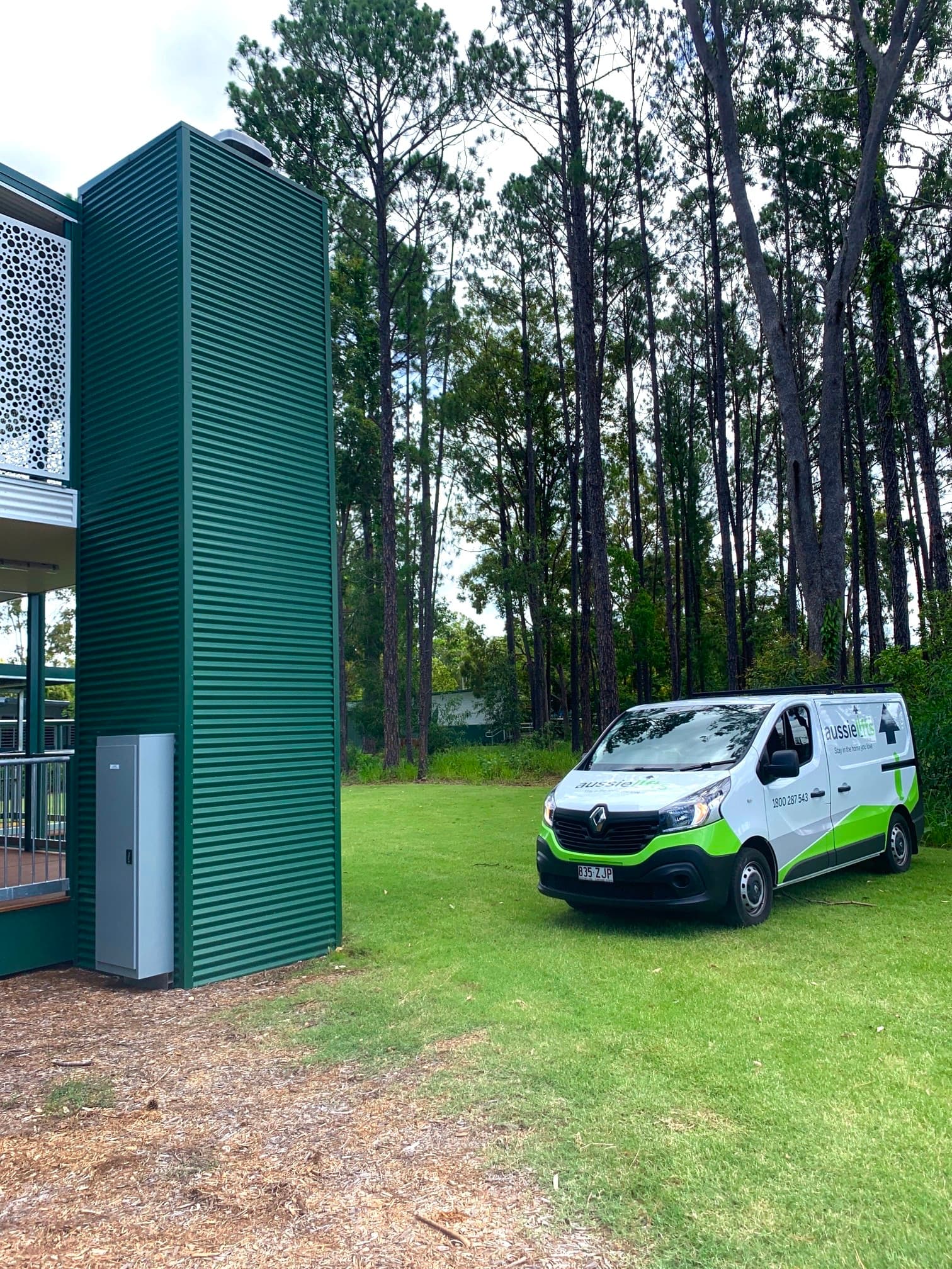 Green clad external lift shaft beside a raised deck, with a white van parked on the lawn under tall pine trees