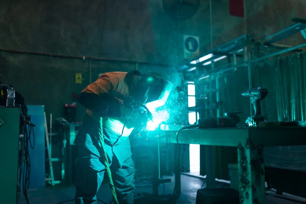 Welder in a helmet working at a steel bench in a fabrication workshop, sparks lighting the scene green