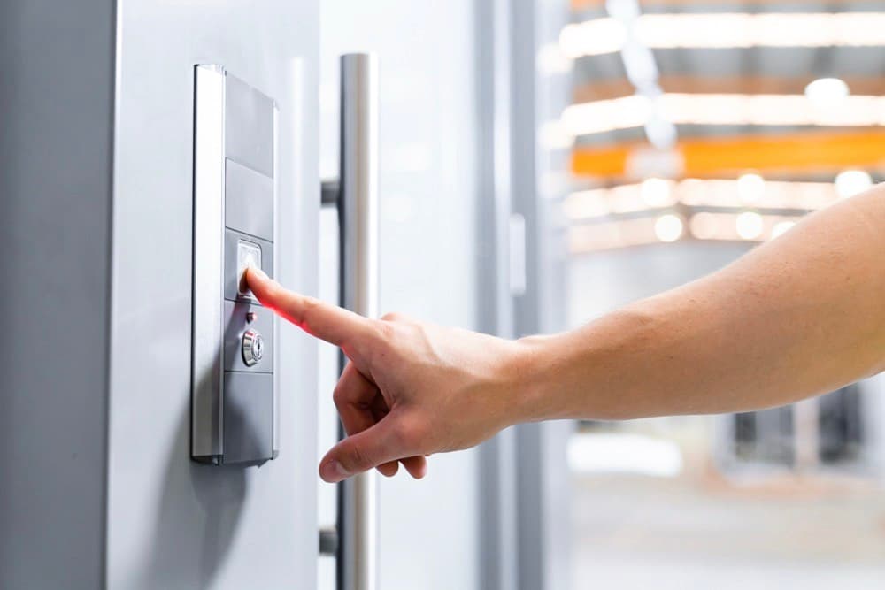 Finger pressing the call button on a stainless steel lift panel beside a glass door