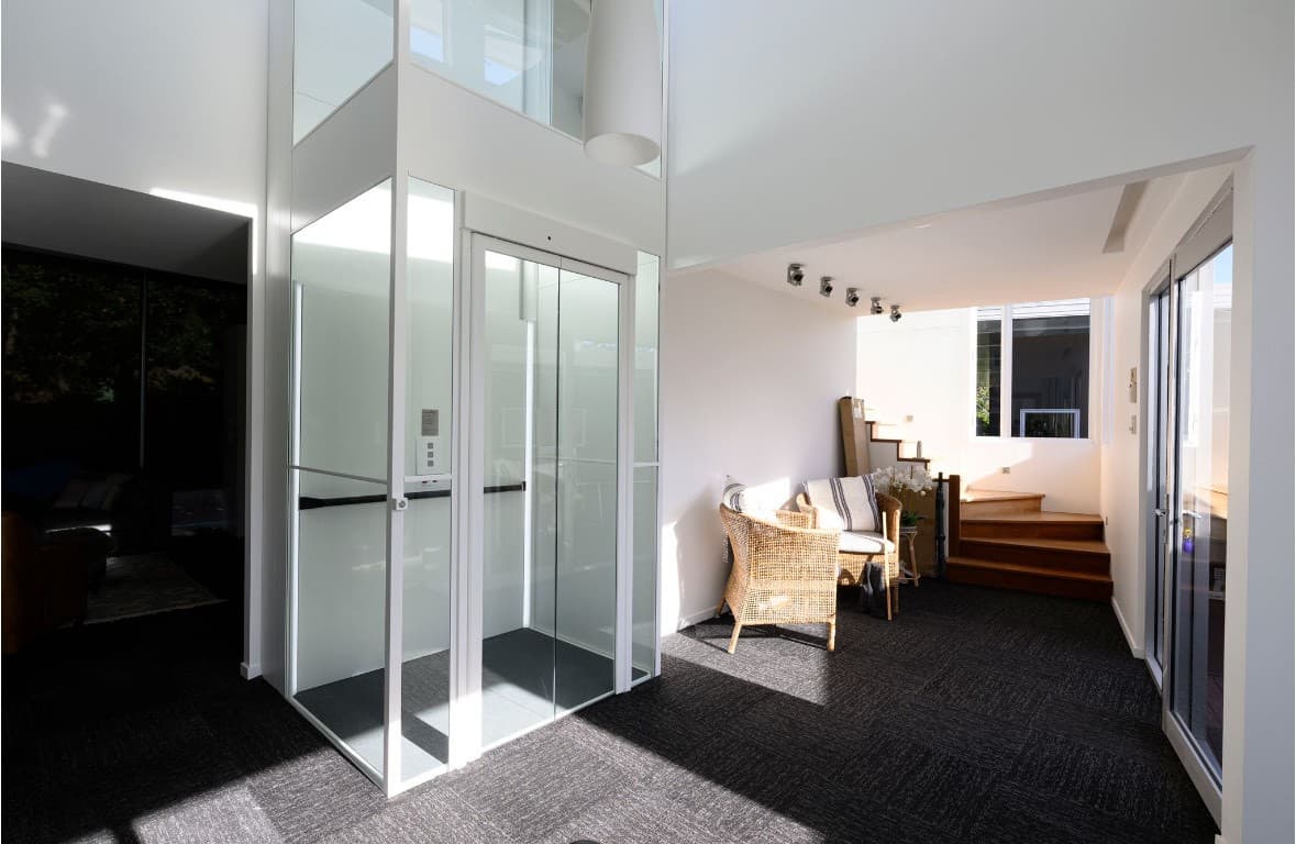 A Swift Lite home lift with a white frame and clear glass shaft and doors, beside a wicker armchair and timber stairs in a Holland Park home