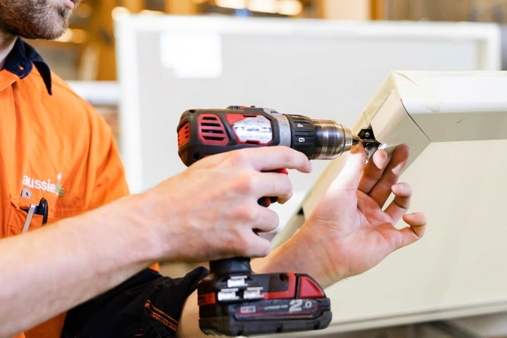 Technician in an orange Aussie Lifts work shirt using a cordless drill on a white lift panel