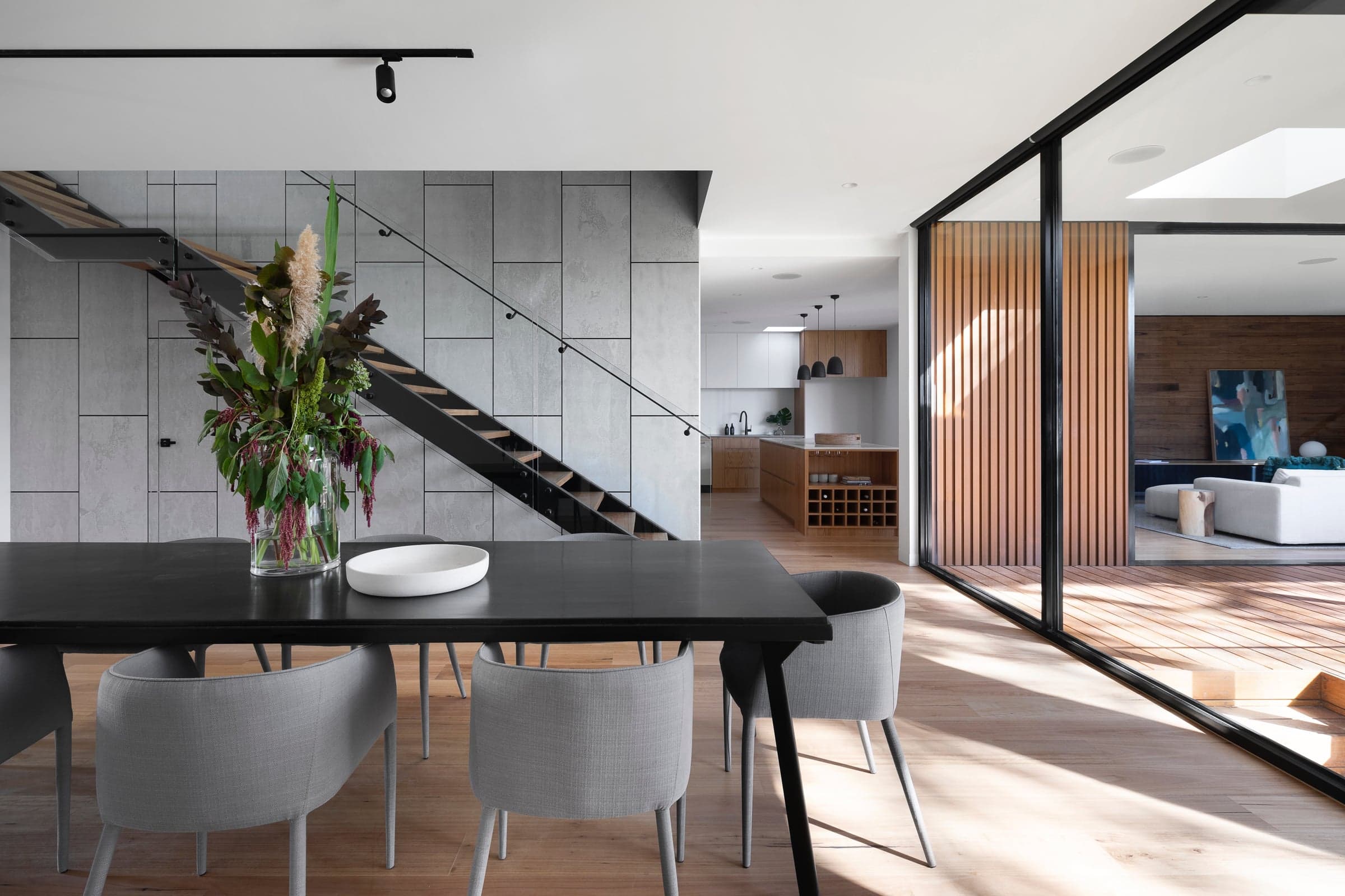Open plan dining room with a black table and grey chairs, a floating black steel staircase against concrete panels and the kitchen beyond