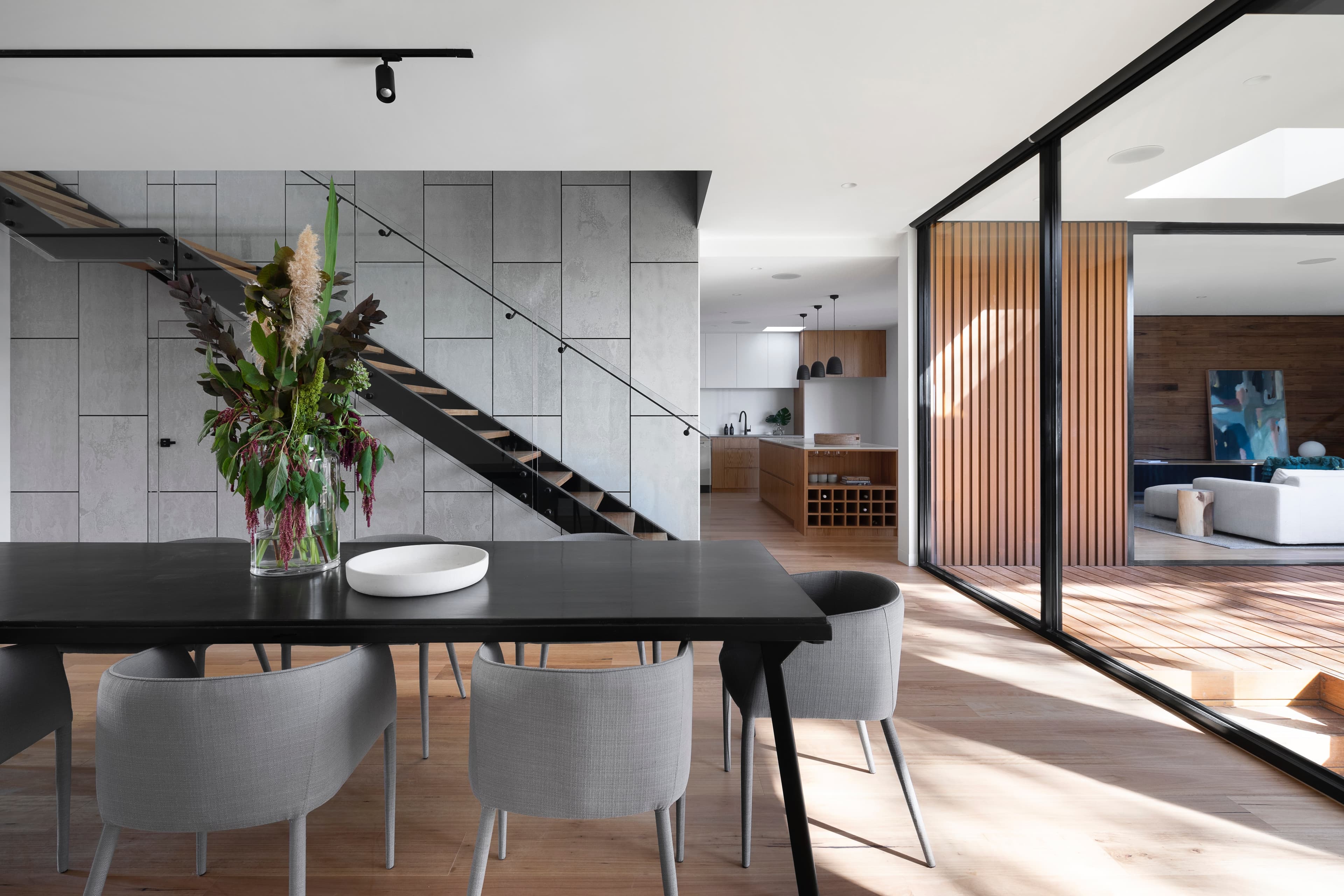 Open plan dining room with a black table and grey chairs, a floating black steel staircase against concrete panels and the kitchen beyond
