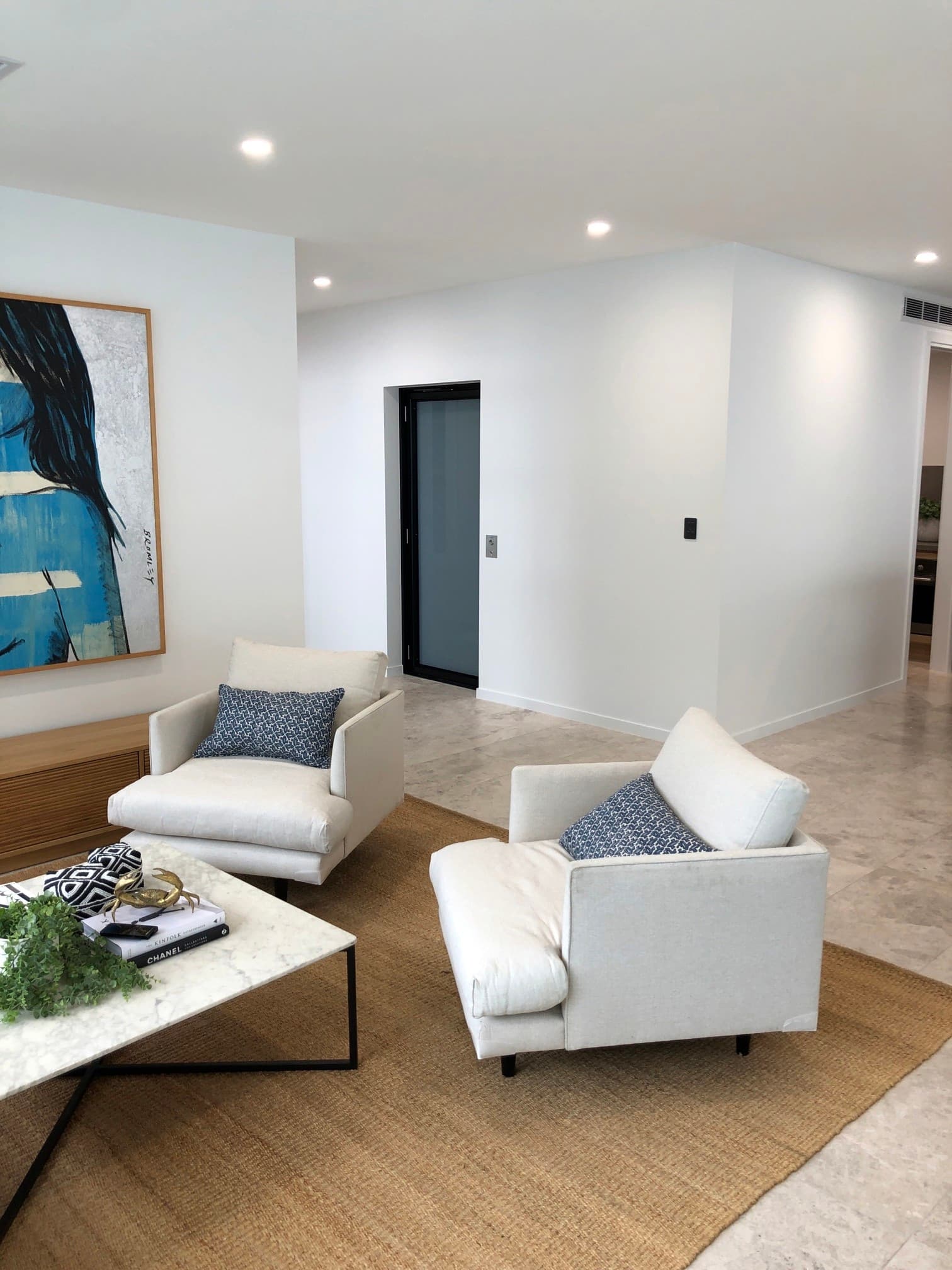 Living room with two white armchairs on a jute rug, and a home lift with a black-framed glass door set into the white wall behind