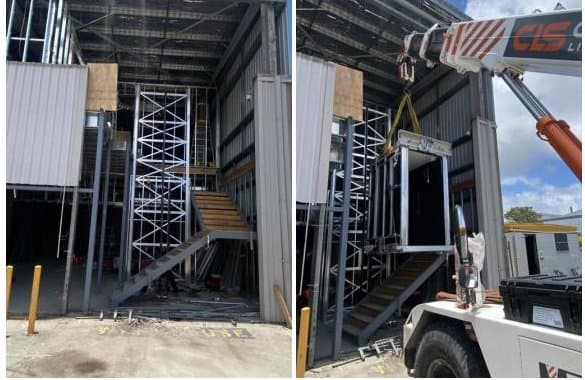 A lift shaft frame standing inside a building under construction, and a crane lowering the second half of the shaft