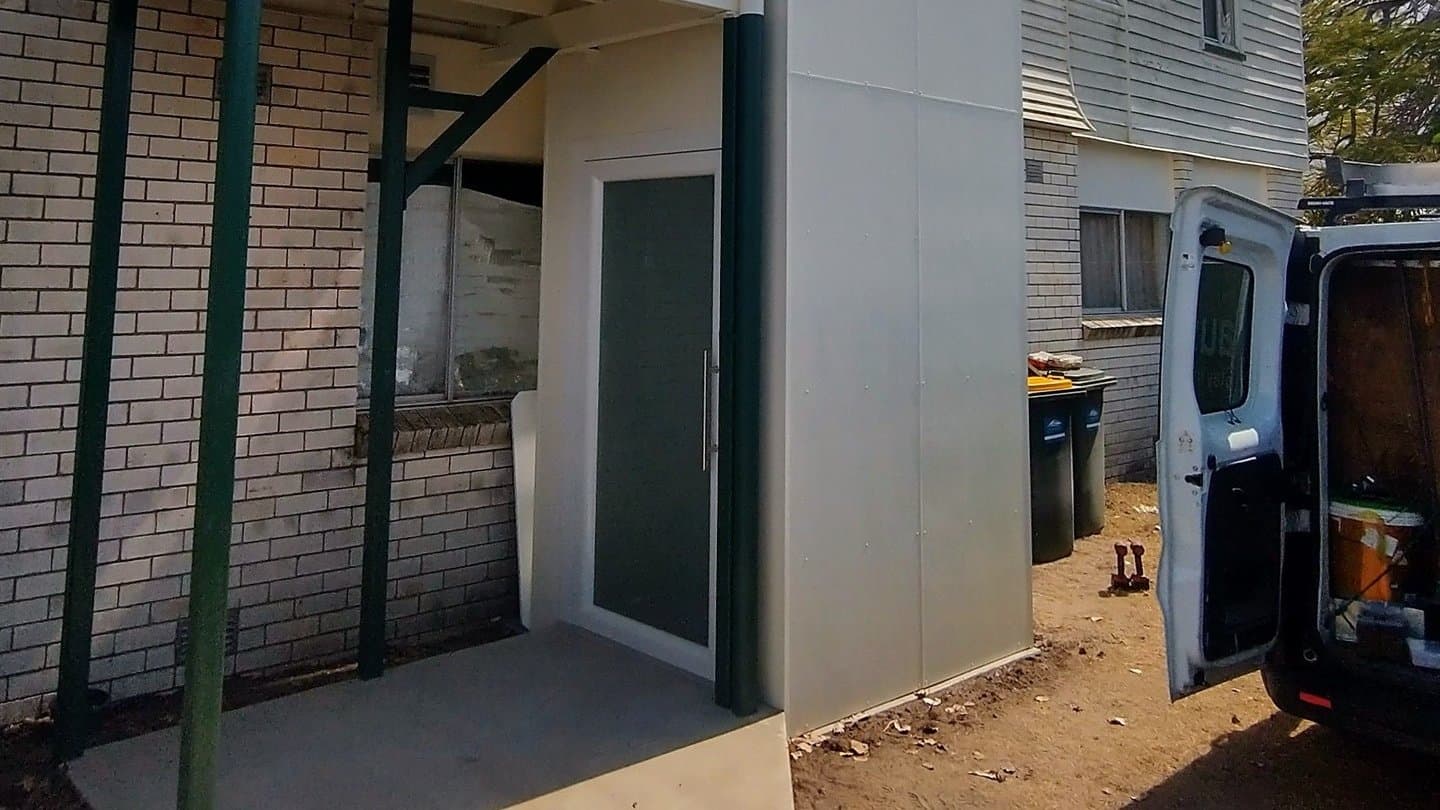 A Phoenix home lift in a Bundaberg, Queensland home, finished white external shaft beside a two-storey house with stairs and a van