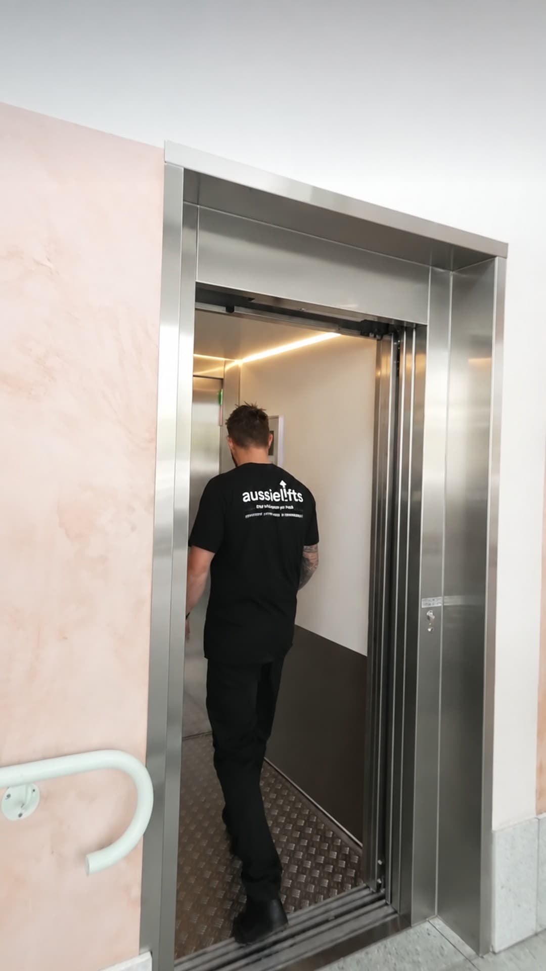 A team member in an Aussie Lifts shirt stepping into the Custom Orion commercial lift at Burleigh Pavilion, Gold Coast, with brushed stainless doors open and cabin lighting visible