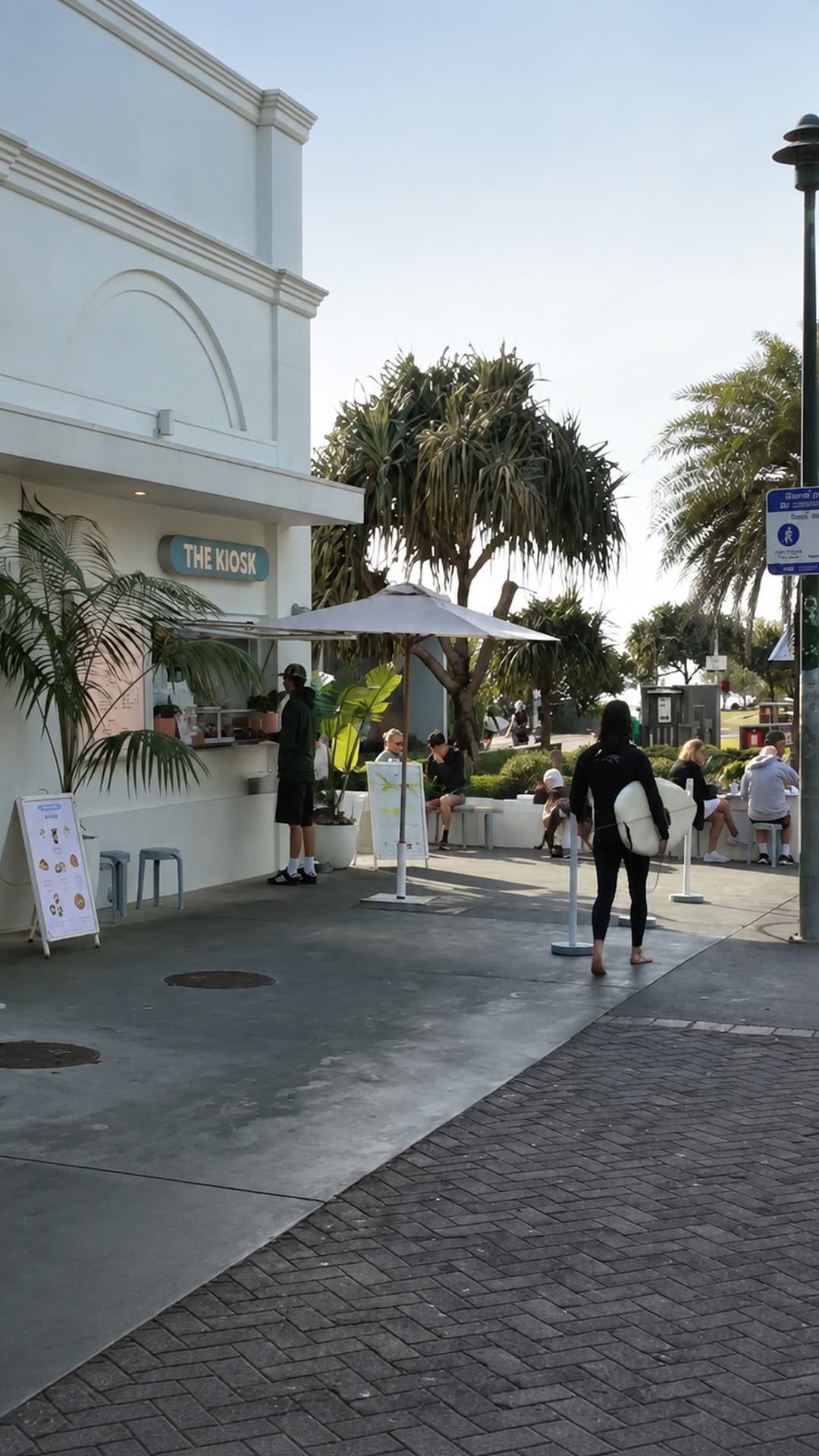 THE KIOSK outdoor service window at Burleigh Pavilion on the Gold Coast, with patio seating, tropical planting and a surfer on the promenade