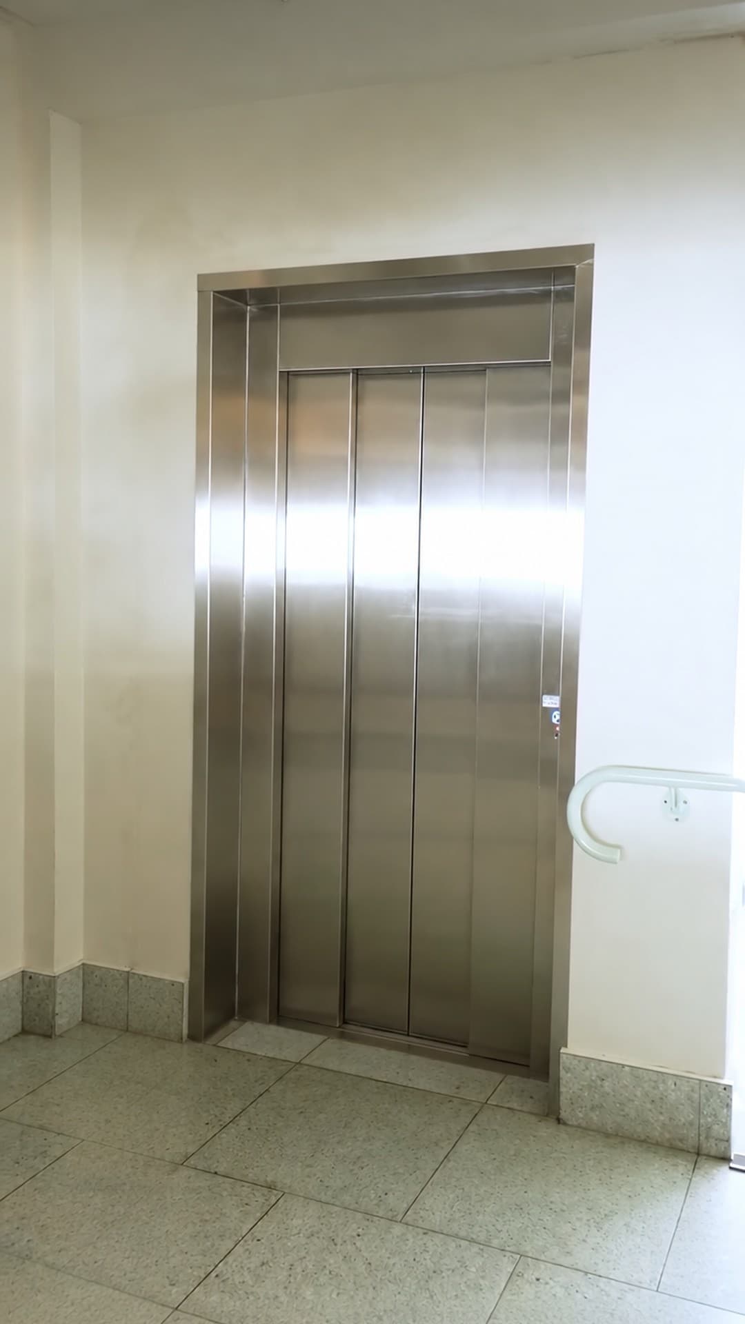 Closed brushed stainless doors of the Custom Orion lift in a bright hallway at Burleigh Pavilion, Gold Coast, with a white handrail beside the landing