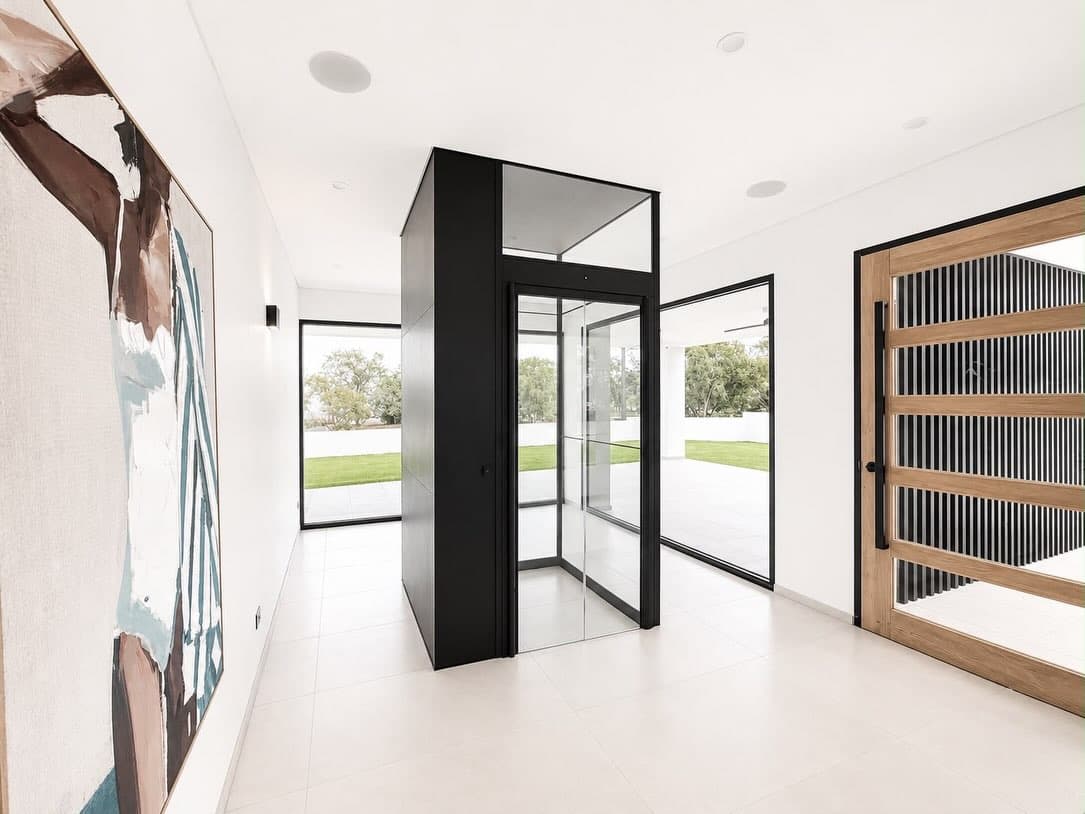 Jet Black Swift Pro glass home lift in the Fannie Bay foyer beside a figurative painting, with a lawn and trees visible through the windows