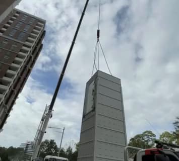 A Phoenix lift being craned into a Macquarie Park, Sydney development by ANC Cranes, the modular shaft hanging from the crane hook beside a residential tower