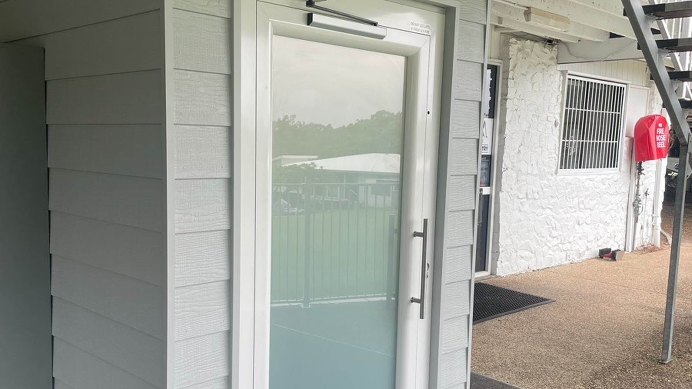 A Phoenix lift at Mt Coolum Golf Course on the Sunshine Coast, white-framed frosted landing door in a grey weatherboard shaft under the clubhouse deck, with a fire hose cabinet nearby