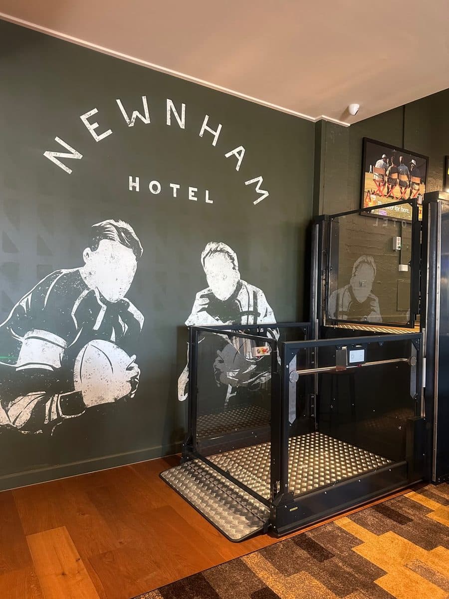 Discovery platform lift with glass panels and an open gate beside the Newnham Hotel mural, Wishart