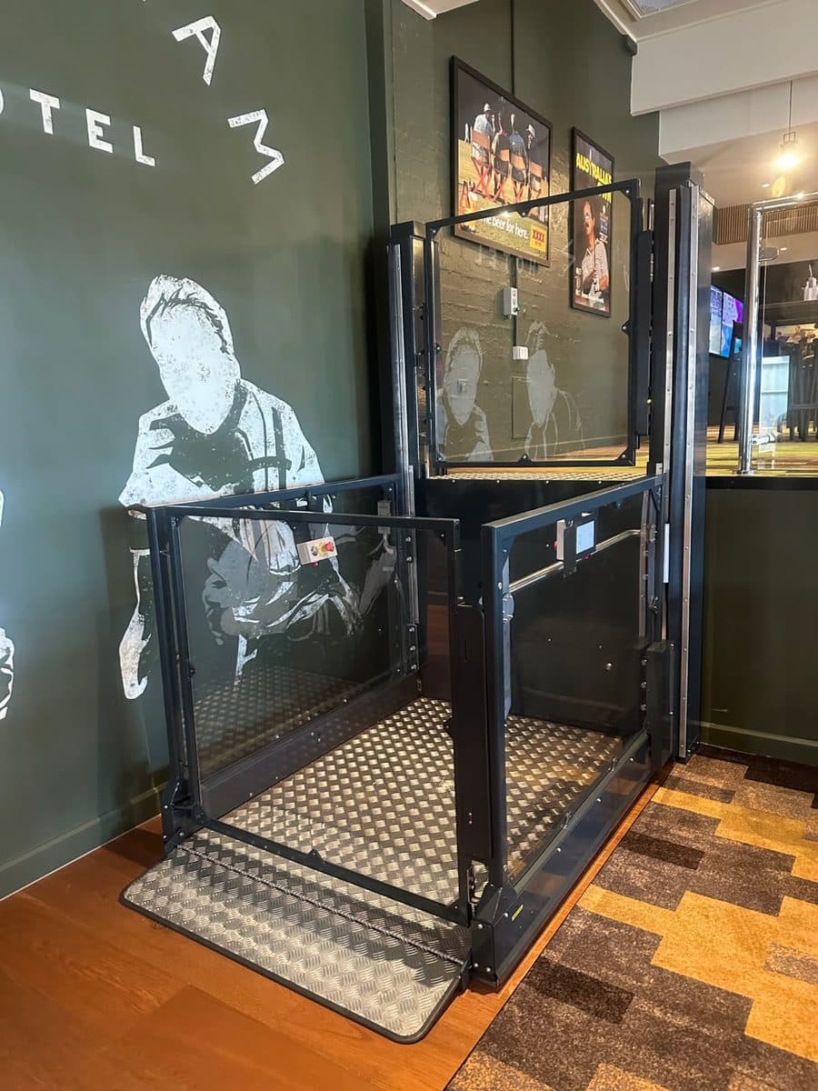 Discovery platform lift with a dark steel frame and glass panels beside the mural, gates open onto the chequer-plate floor, Newnham Hotel, Wishart