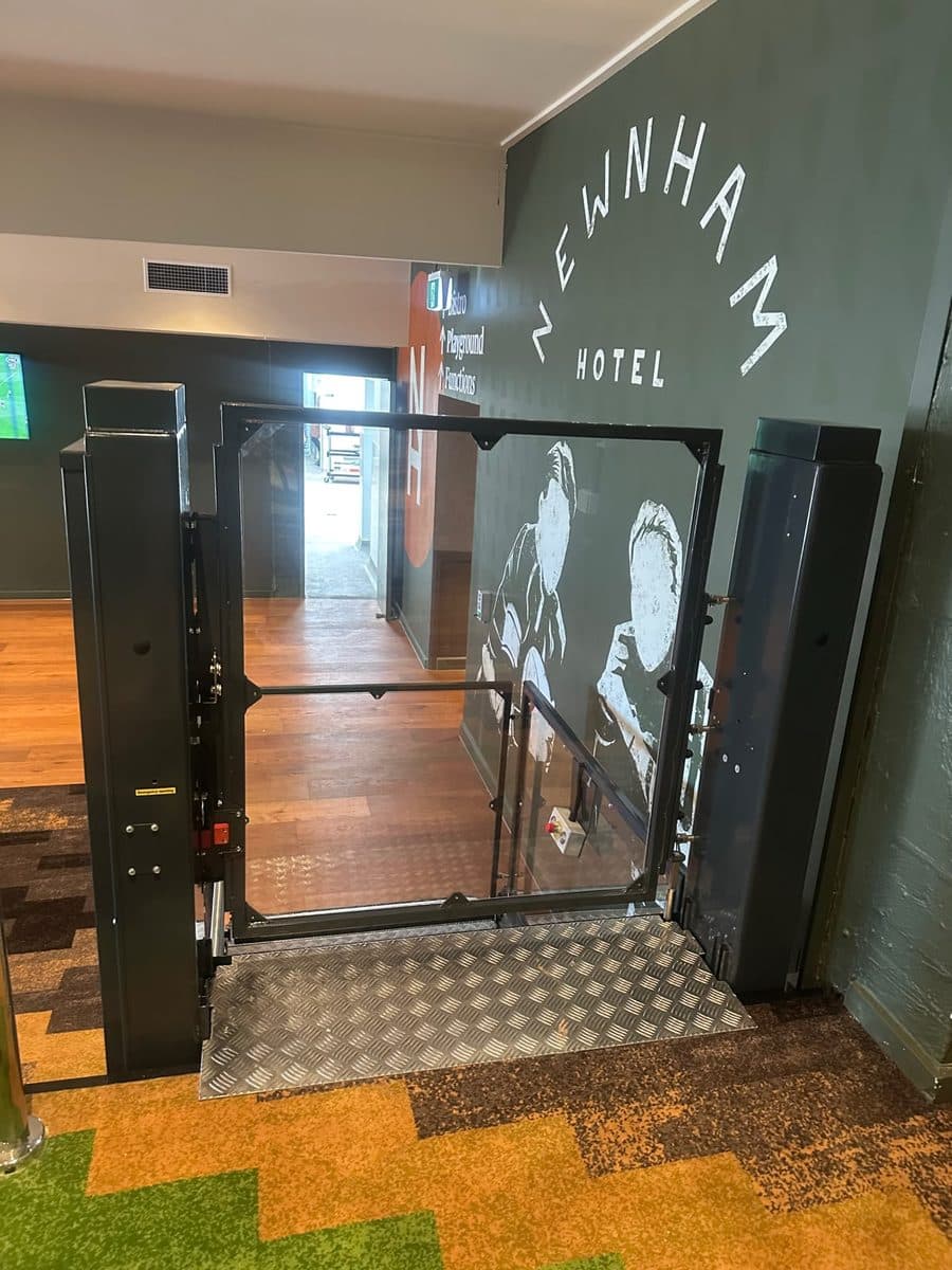 Discovery platform lift's glass gate at the bar level between two dark steel towers, the Newnham Hotel mural beside it, Wishart
