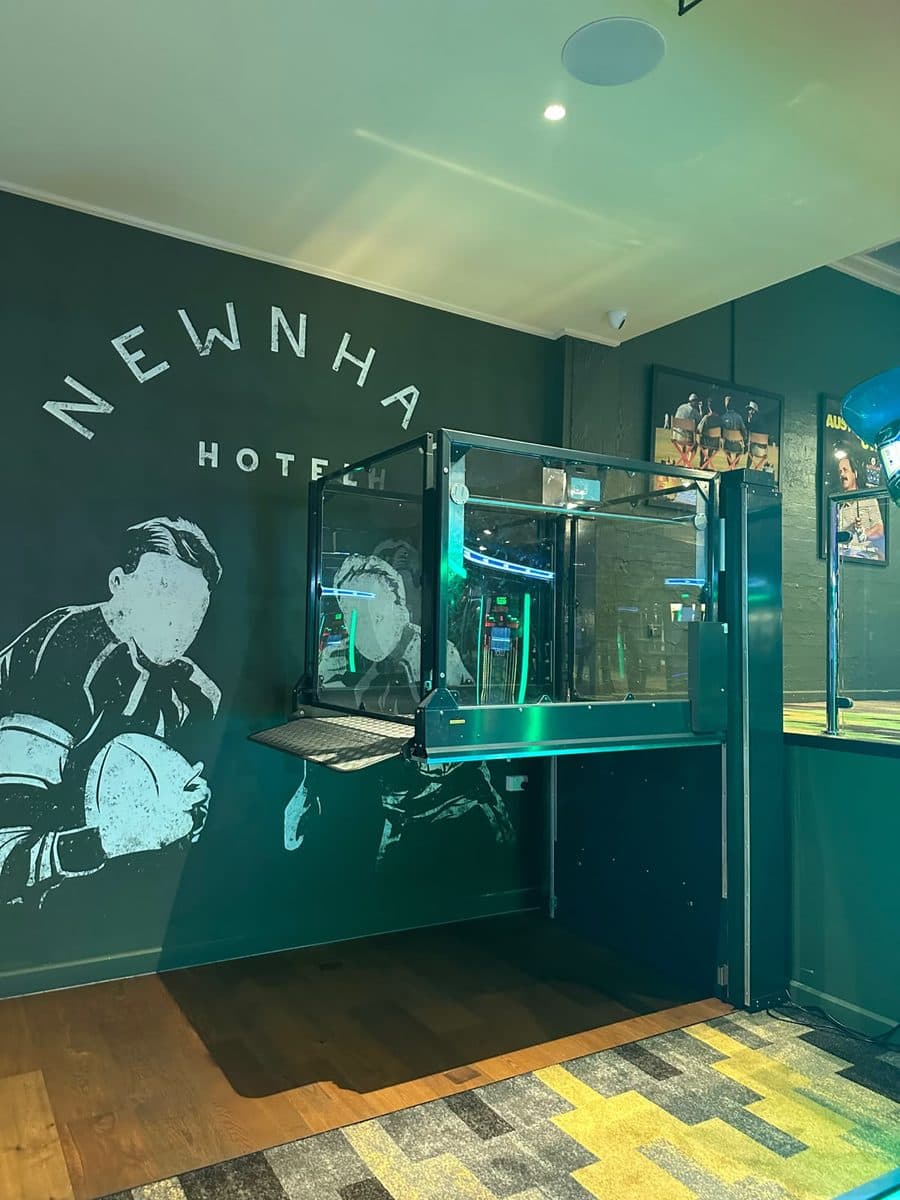 Discovery platform lift raised to the bar level with its ramp down, beside the Newnham Hotel mural under green lighting, Wishart