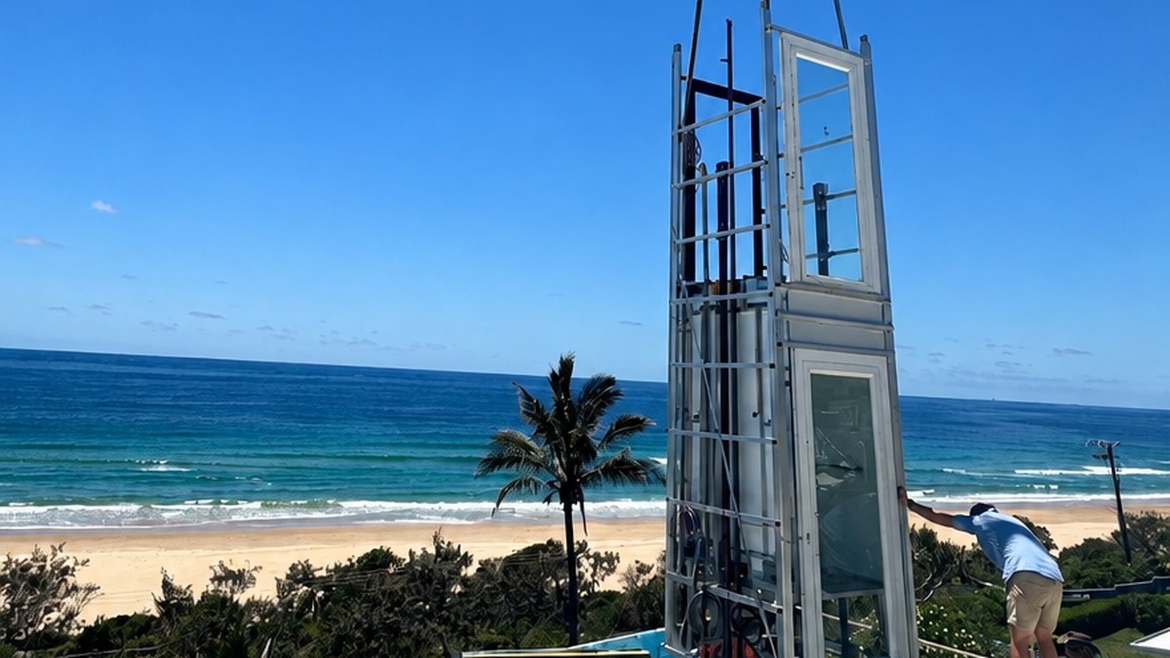 A Phoenix home lift in a beachfront Sunrise Beach home on the Sunshine Coast, craned into place beside a palm tree with the ocean and sandy beach behind