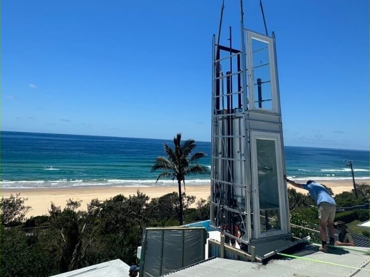 The Phoenix home lift shaft being craned onto the roof of a beachfront Sunrise Beach home on the Sunshine Coast, with the ocean, beach and a palm tree behind