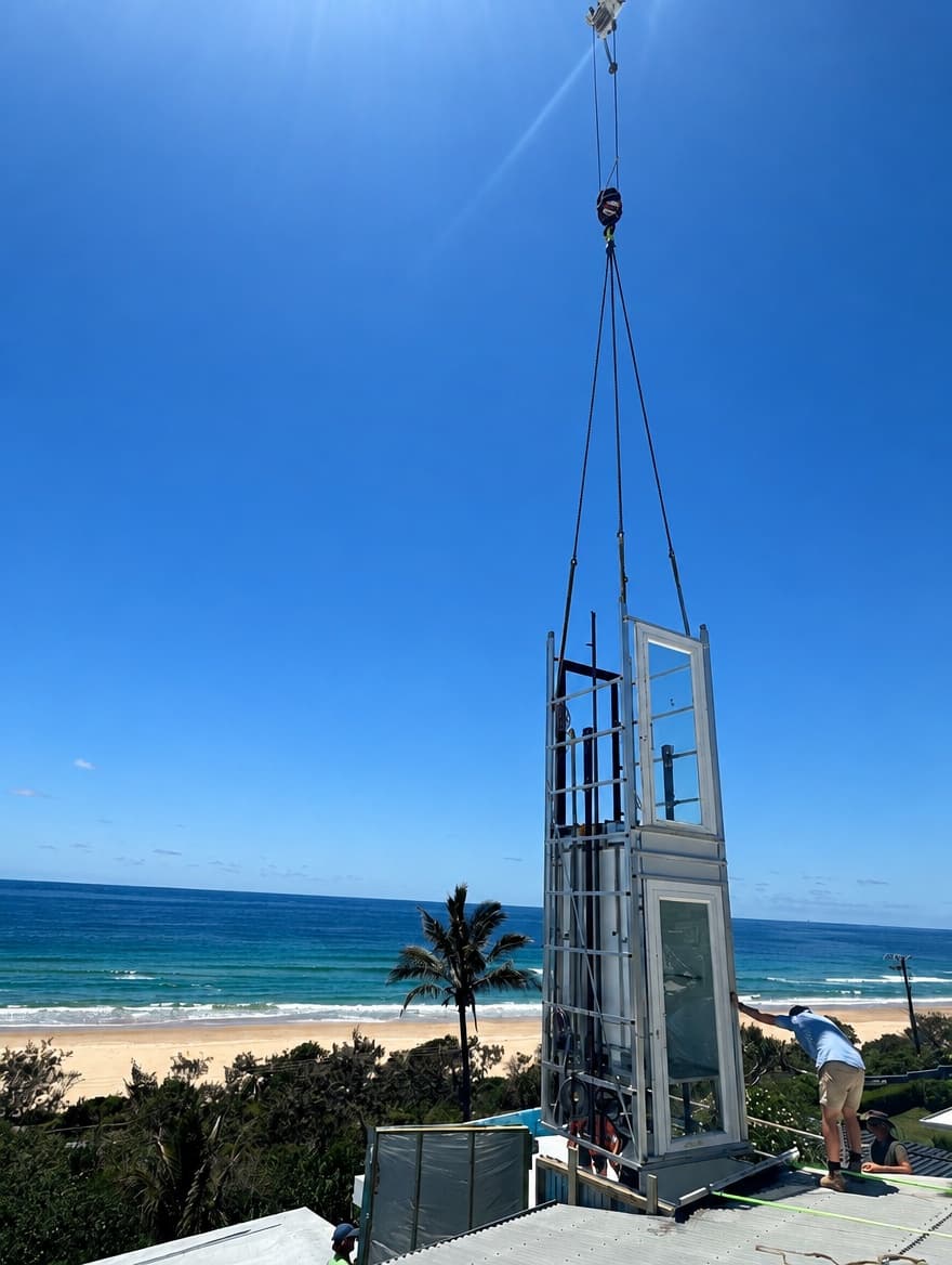 The Phoenix home lift shaft being craned onto the roof of a beachfront Sunrise Beach home on the Sunshine Coast, with the ocean, beach and a palm tree behind
