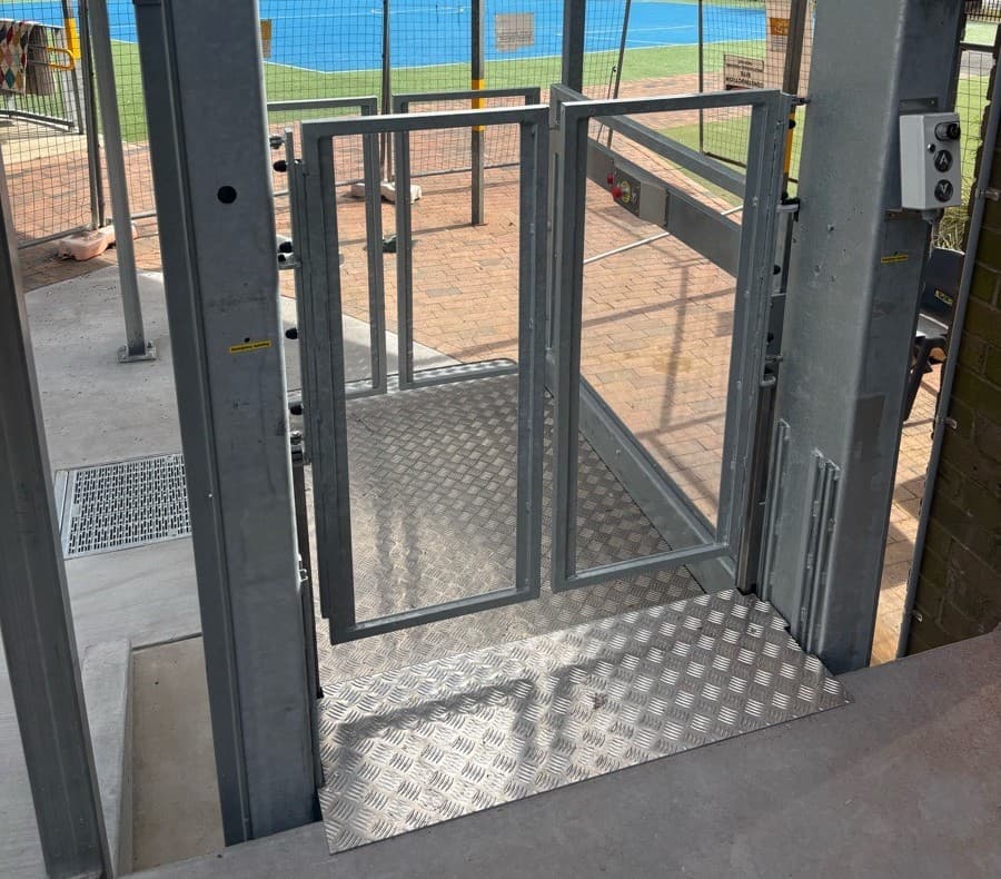 Looking through the open glass gates of the Discovery platform lift at the top landing, its chequer-plate floor and call buttons on the galvanised steel frame, toward the courts