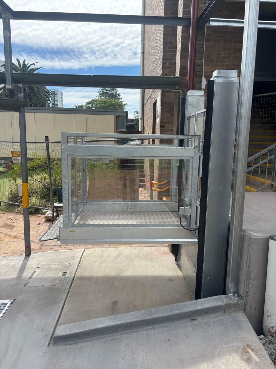 Discovery platform lift at the lower level, a galvanised steel frame with glass panels and a chequer-plate floor beside a brick building and stairs
