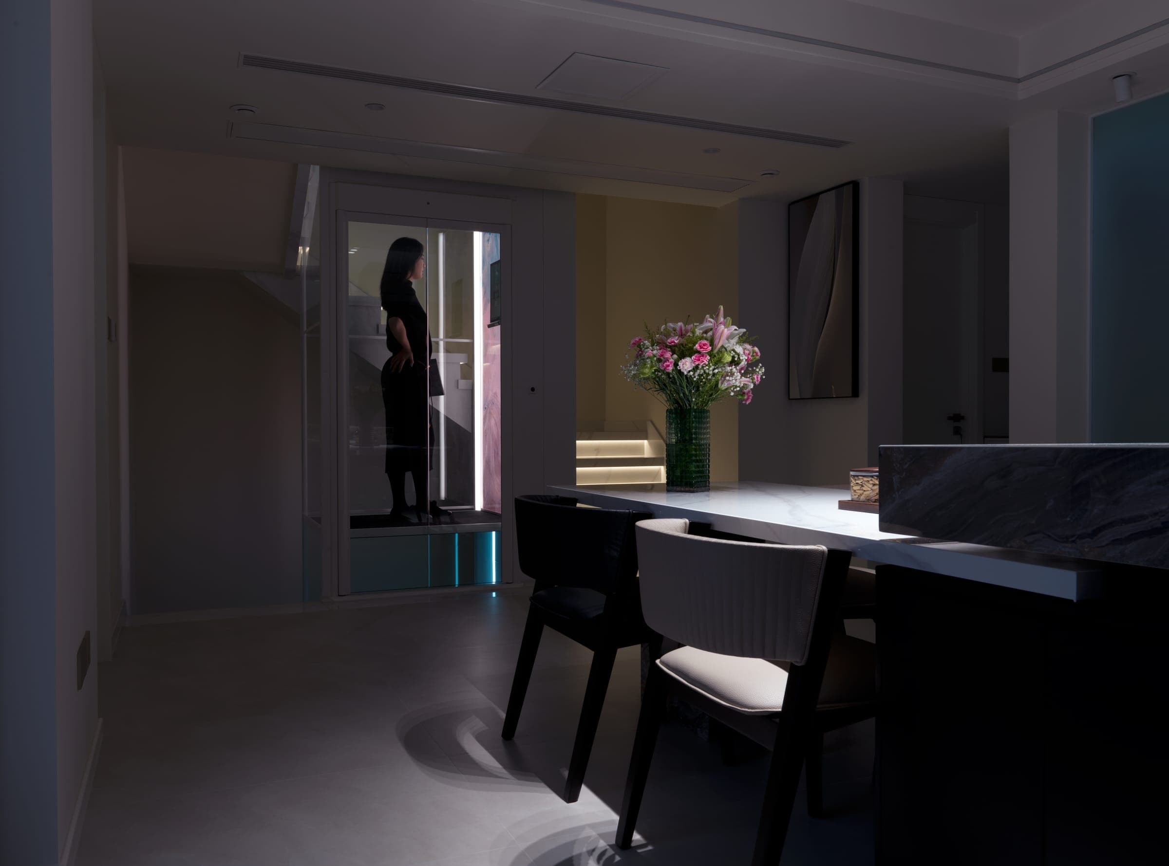 A Swift Pro home lift with its door open in a dimly lit open plan kitchen and dining room, a woman standing inside the lit car